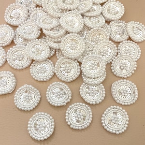 May include: A collection of white, circular embellishments. Each disc features a central design of small, round beads surrounded by a ring of sequins and an outer ring of larger beads. The items are scattered across a neutral surface.