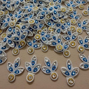 Puede incluir: Primer plano de múltiples adornos florales blancos, azules y dorados. Cada adorno presenta un círculo central dorado rodeado de perlas blancas, gemas azules en forma de lágrima y pedrería transparente. Los adornos están dispersos sobre una superficie.