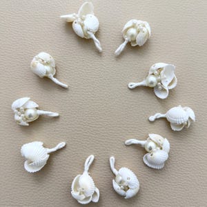 May include: A collection of nine white seashell and pearl embellishments arranged in a circle. Each piece features a small white seashell paired with a central cream-coloured pearl and smaller accent beads, attached to a short white fabric stem.