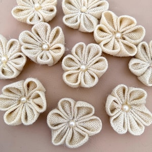 Puede incluir: Un conjunto de diez flores de arpillera beige, cada una con cinco pétalos de tela plegados dispuestos en forma circular. Una pequeña cuenta de perla de color blanco roto, lisa y redonda, está fijada en el centro de cada adorno floral.