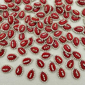 Pode incluir: Uma coleção de pequenos cabochões vermelhos em forma de lágrima, cada um apresentando uma fila vertical central de strass transparentes e uma borda de pequenos cristais claros. Estes ornamentos decorativos estão dispostos num padrão disperso.