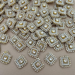 May include: A collection of square decorative embellishments. Each square features a central clear rhinestone, surrounded by small white faux pearls and a gold-coloured border. The embellishments are scattered across a neutral surface.