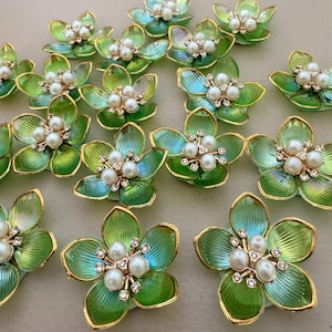 以下が含まれることがあります： 緑と金のフラワーシェイプの装飾品、パールとラインストーンのアクセント付き。 花びらはグラデーション効果があり、緑から青に変化します。 金色のトリムが、これらの装飾的な花の要素にエレガンスを添えています。