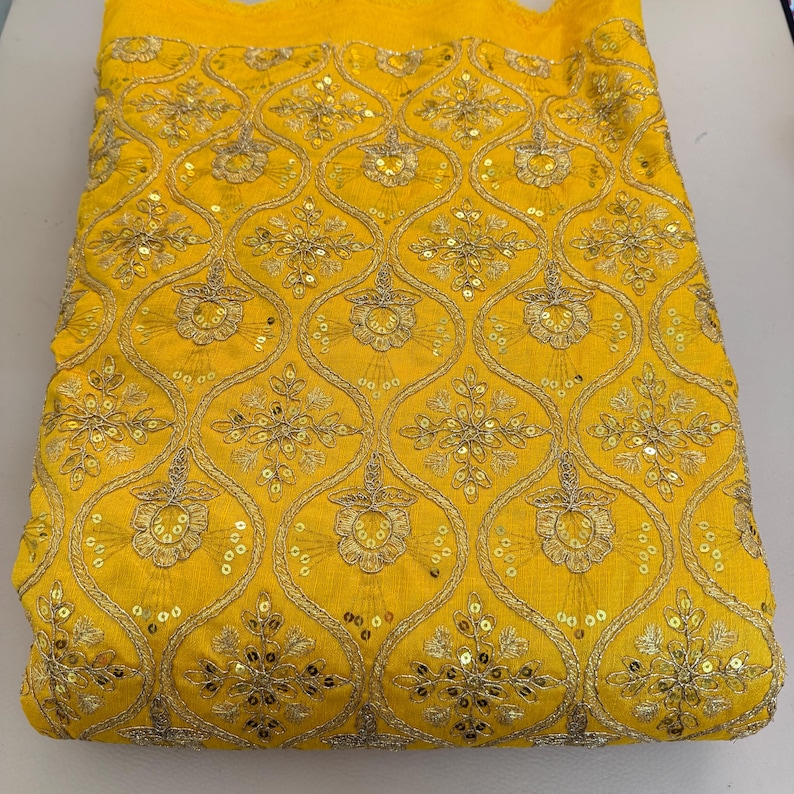 May include: A bright yellow fabric featuring an intricate gold embroidered pattern of repeating ogee shapes. Each section contains a central floral motif accented with small, reflective gold sequins that catch the light across the surface.