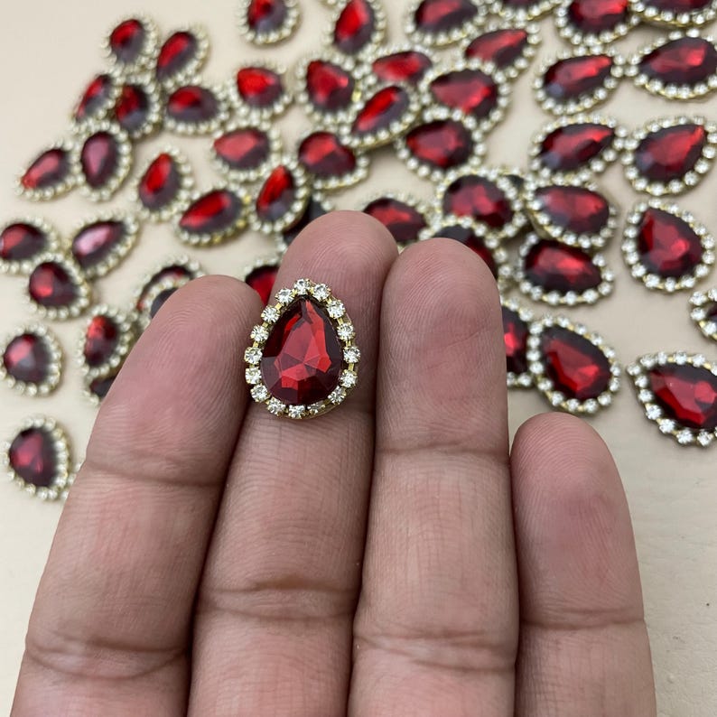 Op de afbeelding: Rode, druppelvormige strass steentjes met een goudkleurige rand en een ring van kleine, heldere strass steentjes. De edelstenen zijn verspreid over een oppervlak, met &eacute;&eacute;n in een hand. Geschikt voor knutselprojecten.