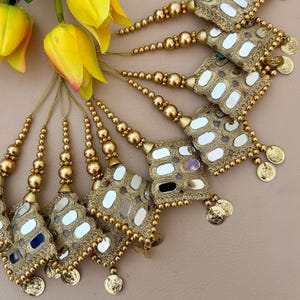 May include: Gold decorative tassels featuring small mirrors and coin-shaped charms. The tassels are arranged in a fan shape, with yellow flowers in the upper left. The tassels have a beaded design, likely for embellishment.