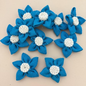 Puede incluir: Un conjunto de ocho flores de cinco pétalos hechas de tela de arpillera azul brillante. Cada flor presenta un centro circular texturizado de color blanco roto hecho de hilo tejido. Los pétalos están dispuestos en forma de estrella, mostrando el tejido grueso del material azul frente a los centros de color neutro.