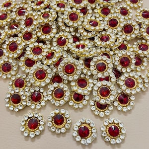May include: A pile of gold-coloured embellishments, each with a central red jewel surrounded by clear rhinestones and a gold border. These circular pieces are suitable for craft projects. Measurements are in centimetres.