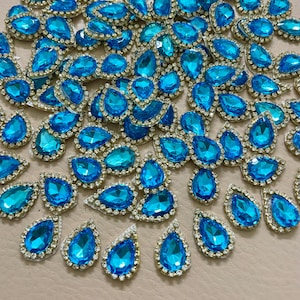 Può includere: Una collezione di strass blu brillante a forma di goccia con un bordo argentato tempestato di strass. Le gemme sono sparse su una superficie dai toni neutri, creando un effetto scintillante. Questi abbellimenti sono ideali per aggiungere brillantezza a oggetti artigianali e accessori.