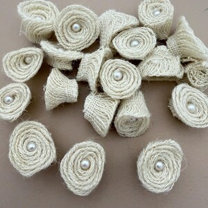 Puede incluir: Una colección de pequeñas rosetas circulares hechas de tela de arpillera texturizada de color blanco roto. Cada adorno individual en forma de flor presenta una cuenta de perla blanca, lisa y redonda, fijada en el centro del diseño en espiral.