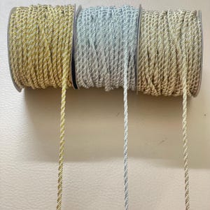 May include: Three spools of decorative twisted cord in gold, silver, and beige. Each spool is tightly wound, with a strand of the cord extending downwards. The cord has a metallic sheen, suitable for crafts and embellishments.