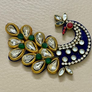 Op de afbeelding: Een decoratieve pauwvormige versiering met een blauwe basis, versierd met heldere, druppelvormige kristallen, kleine groene kralen en goudkleurige afwerking. De kop van de pauw heeft een rode accentkleur en extra kristallen.