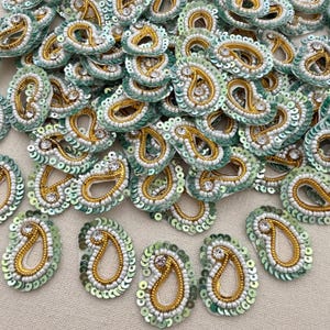 May include: A collection of teardrop-shaped embroidered patches featuring a central gold beaded outline, a white beaded inner border, and a light green sequined edge. Each patch is accented with a single clear crystal at the top point.