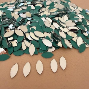 Pode incluir: Uma coleção de peças de vidro espelhado em forma de folha com um suporte verde. As superfícies refletoras do vidro criam um efeito brilhante. Estes elementos decorativos estão espalhados por uma superfície, sugerindo o seu uso em projetos de artesanato ou design.