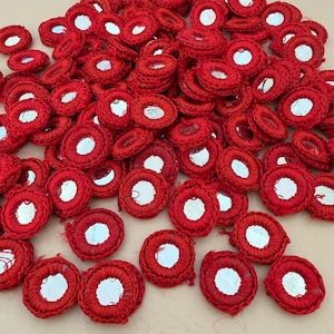 Puede incluir: Un montón de pequeños parches redondos bordados en rojo, con un disco espejado central. Los parches están densamente empaquetados, mostrando el intrincado trabajo de hilo y las superficies reflectantes. Estos elementos decorativos podrían usarse para embellecer ropa o manualidades.