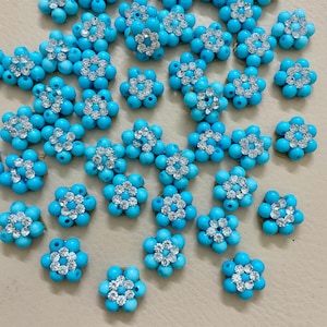 May include: A collection of turquoise-colored flower-shaped beads. Each bead is composed of small, round turquoise beads arranged in a floral pattern, with a silver-colored rhinestone center. The beads are scattered on a light beige surface.