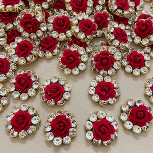 Op de afbeelding: Close-up van meerdere decoratieve versieringen. Elke versiering heeft een kleine rode roos in het midden, omringd door heldere, sprankelende strass-steentjes en een goudkleurige rand. Deze worden waarschijnlijk gebruikt voor knutselen of om details aan kleding toe te voegen.