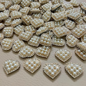 Pode incluir: Uma coleção de enfeites em forma de coração. Cada coração apresenta uma moldura de metal dourado, preenchida com pérolas falsas e contornada com pequenos strass transparentes. Os enfeites estão espalhados pela superfície.