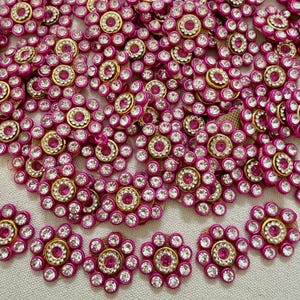 Könnte beinhalten: Eine Sammlung blumenförmiger Verzierungen mit einem zentralen rosa Strassstein, umgeben von einem Ring aus klaren weißen Strasssteinen, alles in einem goldfarbenen Metallrahmen mit einem leuchtend rosa Außenrand aus kleinen runden Akzenten.
