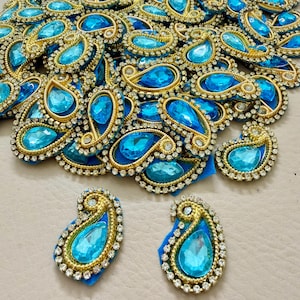 May include: A collection of ornate, teardrop-shaped embellishments. Each piece features a large, light blue gemstone set within a gold-colored frame, surrounded by small, clear crystals. The base is a deep blue fabric.