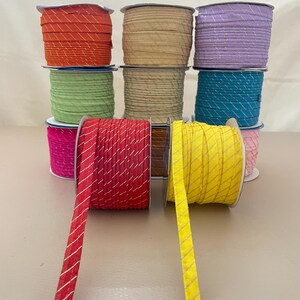 Pode incluir: Uma coleção de rolos de fita decorativa em várias cores, incluindo laranja, bege, lilás, turquesa, verde, vermelho, amarelo e rosa. Cada fita apresenta um padrão de listras diagonais douradas. Os rolos estão empilhados.