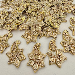 May include: A selection of gold-toned, star-shaped embellishments with a paisley design. Each piece is decorated with small, sparkling, amber-coloured rhinestones. These embellishments are likely for crafts or adding decorative detail to clothing or accessories.