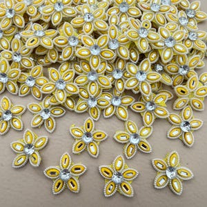 May include: A collection of yellow flower-shaped appliqués featuring five petals each. Every petal contains a small rectangular clear crystal, and a larger round clear crystal is positioned in the centre of each flower. The edges are outlined in white.