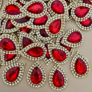 May include: A collection of teardrop-shaped embellishments featuring a central red faceted stone surrounded by a border of small clear rhinestones set in a gold-toned metal frame. Each piece is individually crafted with a pointed top and rounded base.