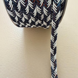 May include: A spool of decorative trim with a braided design. The trim features a pattern of navy blue, white, and brown segments, creating a chevron-like appearance. The trim is tightly wound on a dark brown spool.
