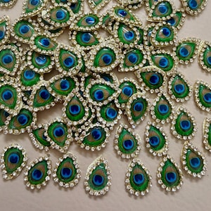May include: A collection of decorative peacock feather embellishments. Each teardrop-shaped piece features a vibrant blue and green peacock eye design, outlined with a gold border and encrusted with small, clear rhinestones. The pieces are scattered across a neutral surface.