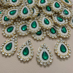 May include: A collection of teardrop-shaped embellishments. Each piece features a large, emerald-green gemstone at the center, surrounded by a border of small, clear rhinestones and white pearls. The outer edge is trimmed with gold-colored metal.