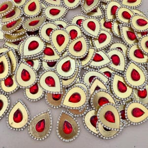 Puede incluir: Una colección de adornos en forma de lágrima con una piedra roja central rodeada por un marco de metal dorado y un borde de pequeños diamantes de imitación transparentes. Cada pieza tiene una parte superior puntiaguda y una base redondeada.
