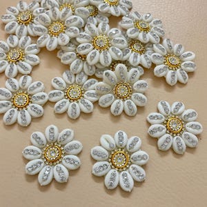 Può includere: Una collezione di abbellimenti bianchi a forma di margherita con dettagli dorati e argentati. Ogni fiore ha un centro dorato circondato da strass trasparenti e perline bianche a forma di petalo. Ideale per progetti di artigianato.