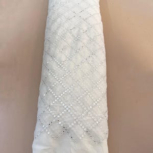 May include: A roll of white fabric with a geometric pattern of small, reflective sequins. The fabric is tightly rolled, showcasing the intricate design and texture. The background is a solid, neutral color.