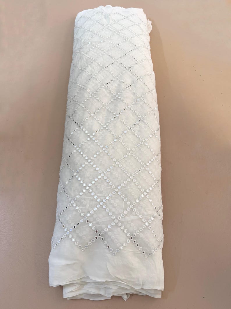 May include: A roll of white fabric with a geometric pattern of small, reflective sequins. The fabric is tightly rolled, showcasing the intricate design and texture. The background is a solid, neutral color.