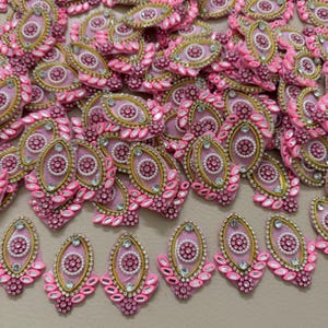 Könnte beinhalten: Eine Ansammlung von ovalförmigen, rosa Stoffverzierungen. Jedes Stück hat ein zentrales Design mit weißen Perlen, umgeben von goldenen Verzierungen und klaren, funkelnden Akzenten. Die Ränder sind mit rosa, tropfenförmigen Verzierungen verziert.