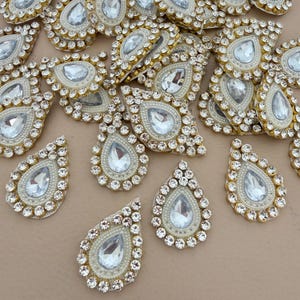 Op de afbeelding: Een verzameling druppelvormige versieringen. Elk stuk heeft een grote, heldere, facetgeslepen edelsteen in het midden, omgeven door een rand van kleine, heldere strass-steentjes en imitatieparels. De randen zijn afgewerkt met goudkleurig materiaal.