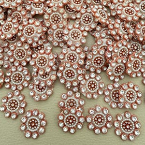 Peut inclure: Une collection de boutons circulaires présentant un groupe central de petites perles blanches entouré d'un anneau de strass transparents à facettes, sertis dans un cadre en métal cuivré. Chaque bouton a un bord festonné avec des strass.