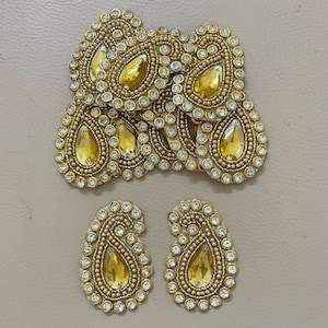 May include: Gold-toned paisley-shaped embellishments with yellow teardrop-shaped center stones and clear rhinestone borders. The embellishments are arranged on a neutral-colored surface. These decorative pieces are suitable for various craft projects.