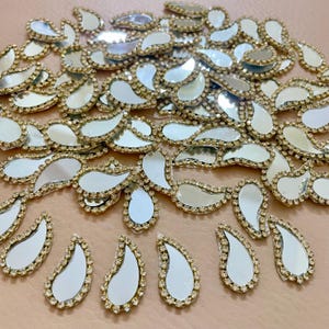 Puede incluir: Una colección de adornos en forma de lágrima con centros espejados y bordes de pedrería dorados. Estas piezas decorativas están dispersas sobre una superficie marrón claro, reflejando la luz.