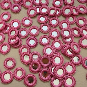 May include: A close-up of numerous small, circular, pink embroidered patches with mirrored centres. The patches are closely packed together, displaying a repeating pattern of circular designs. The pink embroidery surrounds the mirrored centres, creating a decorative effect.