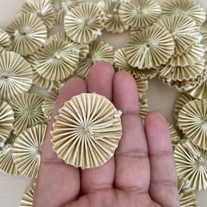 May include: A collection of beige, pleated fabric rosettes. The rosettes are circular with a radial design, resembling small fans. The image shows several rosettes, with one held in a hand, highlighting the texture and color of the decorative elements.