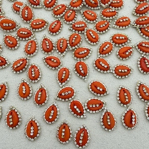 May include: A collection of small, teardrop-shaped orange cabochons, each featuring a central vertical row of three clear rhinestones and a metallic border. These decorative embellishments are arranged in a scattered pattern on a textured surface.