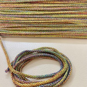 Puede incluir: Un carrete de cordón trenzado colorido en una variedad de colores, incluyendo amarillo, verde, azul, rojo y morado. El cordón está tejido apretadamente y parece estar hecho de un material brillante y metálico. Una porción del cordón está enrollada.
