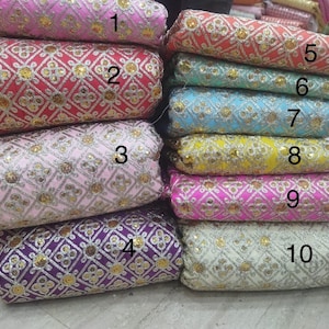 Może przedstawiać: Stos dziesięciu rolek materiału w różnych kolorach, w tym różowym, czerwonym, niebieskim, żółtym i fioletowym. Każda rolka ma haftowany wzór ze złotymi i srebrnymi akcentami. Tkanina ma teksturowany wygląd.