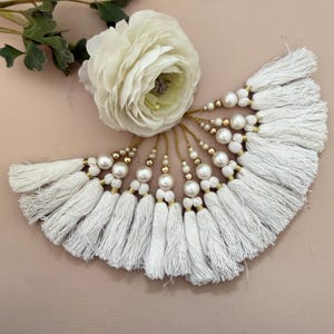 Op de afbeelding: Een verzameling witte stoffen kwastjes gerangschikt in een waaiervorm, elk voorzien van een goudkleurige draadlus, een kleine gouden kraal en een grotere witte kunstparel. De kwastjes liggen bij een witte bloem met groene bladeren.