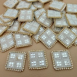 May include: A selection of square embellishments, each with a central design of four smaller squares. They are bordered by small white beads and a gold-coloured rhinestone trim. The embellishments are scattered across a neutral surface, suitable for craft projects.