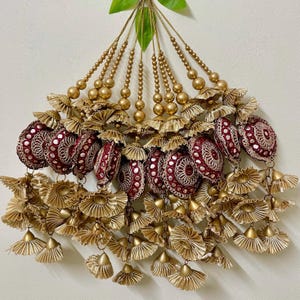 May include: Ornate decorative tassels in gold and burgundy. The design includes gold beaded strands, ruffled gold fabric, and burgundy circular ornaments with silver accents. These tassels are suitable for embellishing various items, with a detailed and intricate design.