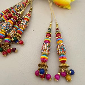 May include: Colorful beaded tassels with gold accents. The tassels feature a variety of vibrant colors, including red, blue, yellow, and green, with a gold bell-shaped base and dangling beads. The beads are arranged in a decorative pattern.