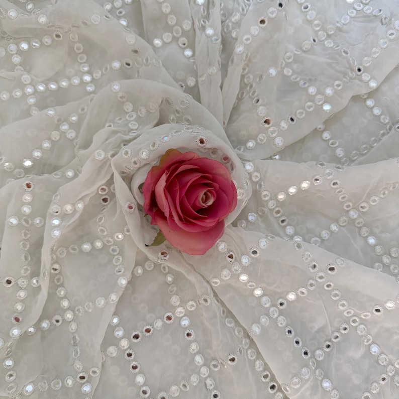May include: White fabric with a geometric pattern of small, reflective circles. A pink rose with green leaves rests on the fabric, adding a pop of color. The fabric is draped, creating soft folds and shadows.